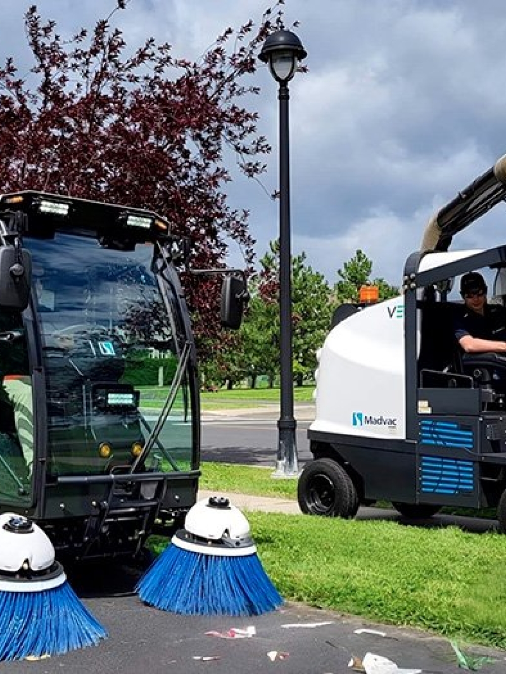 Deux véhicules municipaux de nettoyage, dont une balayeuse et une machine d’aspiration, en opération sur une piste cyclable bordée d’arbres.