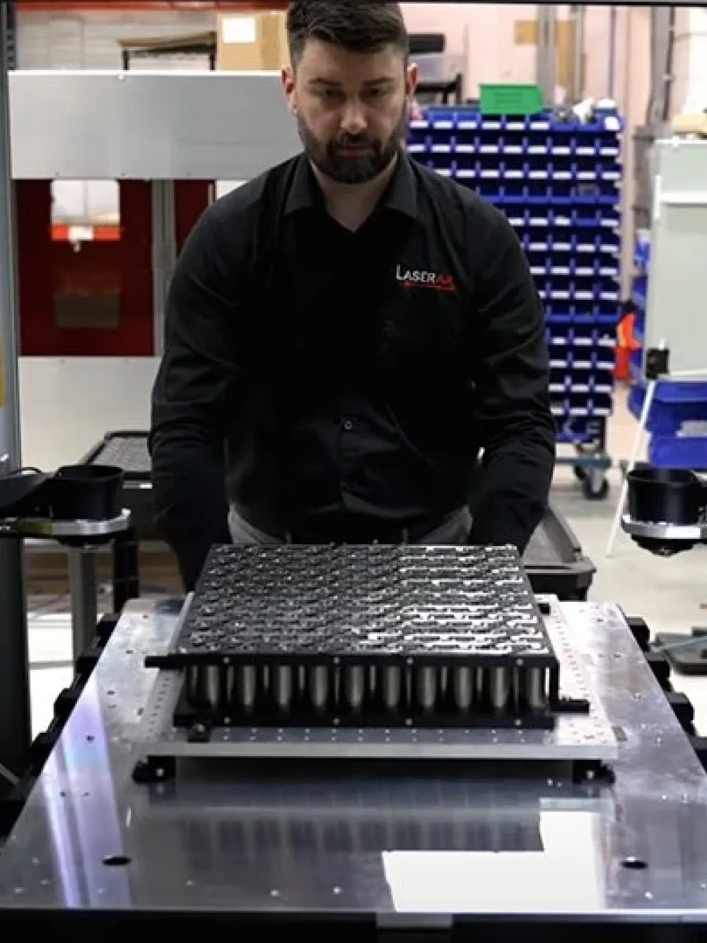 Personne opérant un équipement industriel automatisé avec plusieurs bras robotiques jaunes autour d’une pièce métallique. 