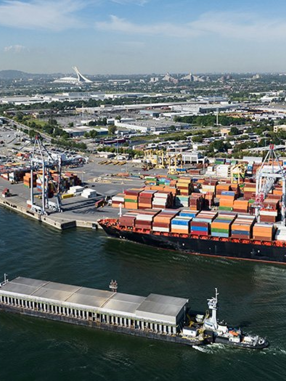 Vue aérienne d’un port avec des navires de marchandises et de nombreux conteneurs colorés, entouré d’infrastructures industrielles et de la ville en arrière-plan.