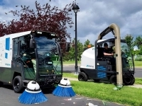 Deux véhicules municipaux de nettoyage, dont une balayeuse et une machine d’aspiration, en opération sur une piste cyclable bordée d’arbres. 