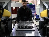 Personne opérant un équipement industriel automatisé avec plusieurs bras robotiques jaunes autour d’une pièce métallique. 