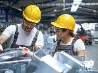 Deux personnes portant des casques de sécurité jaunes travaillent avec des pièces métalliques dans un atelier industriel. 