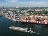 Vue aérienne d’un port avec des navires de marchandises et de nombreux conteneurs colorés, entouré d’infrastructures industrielles et de la ville en arrière-plan. 